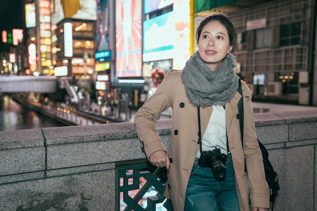 joven viajera con abrigo y bufanda apoyada en el puente en la ciudad nocturna con cartelera en segundo plano. foto al aire libre de una atractiva turista sonriente que se divierte explorando atracciones en osaka, japón.