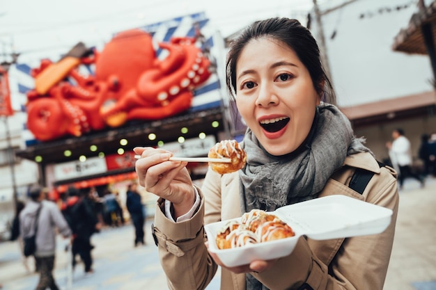 joven viajera asiática probando la cámara facial takoyaki mostrando con palillos. turista emocionado boca abierta comiendo sabrosas bolas de pulpo. deliciosa comida callejera japonesa.