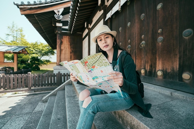 Joven viajera con mochila y sombrero mirando un mapa de papel en el templo shitennoji osaka, japón. Viajar a asia en vacaciones de verano. chica visitante relajada en el santuario sentada en las escaleras de roca pensando.