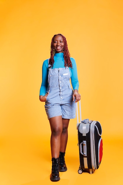 Joven viajera posando con carro, modelo femenina preparándose para salir de viaje de vacaciones sobre fondo naranja. Mujer afroamericana sonriendo y sintiéndose feliz en la aventura urbana.