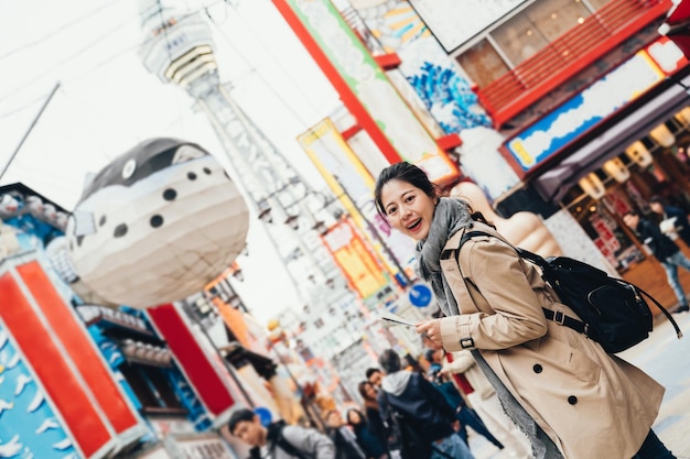 joven viajera sosteniendo un mapa de papel buscando dirección de pie en la concurrida calle en osaka, japón. mochilero femenino sonriendo a la cámara con pez globo y tsutenkaku en segundo plano en un día soleado.