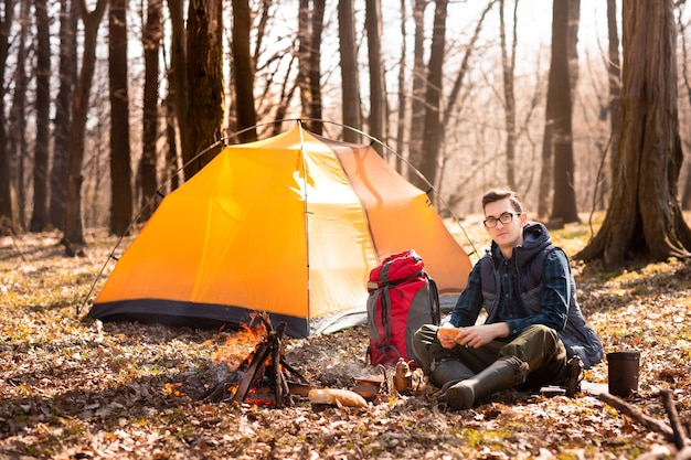 Un joven viajero en el bosque está descansando cerca de la tienda y desayunando en la naturaleza.