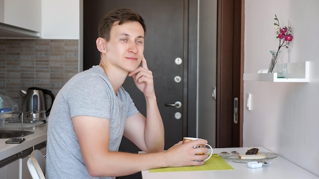 El joven viajero está desayunando. Está sentado a la mesa y bebiendo té. Vista lateral. Está en una pequeña cocina en un piso alquilado de vacaciones.