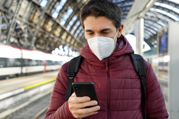 Joven viajero con mascarilla KN95 FFP2 en la estación de tren Joven caucásico usando un teléfono inteligente en la estación de tren