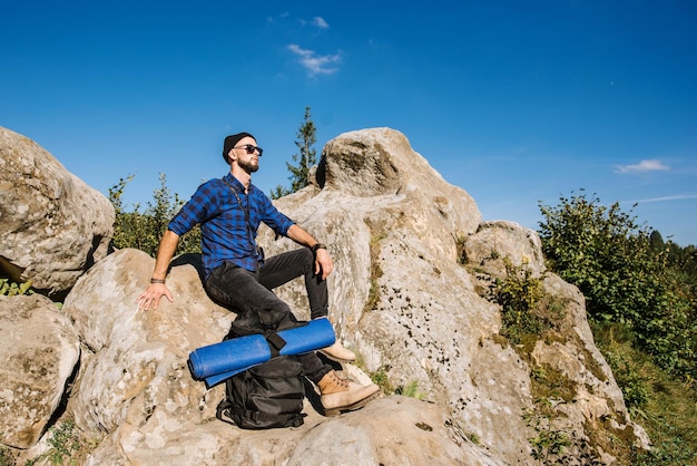 Un joven viajero con mochila con gafas de sol sentado en la cima de las montañas rocosas