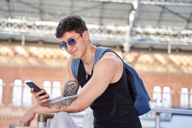 Joven viajero con mochila y gafas de sol usando su teléfono inteligente en la estación