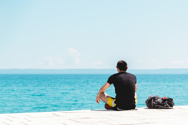 Joven viajero solo con mochila cerca de él meditando mirando hacia el océano y disfrutando del silencio