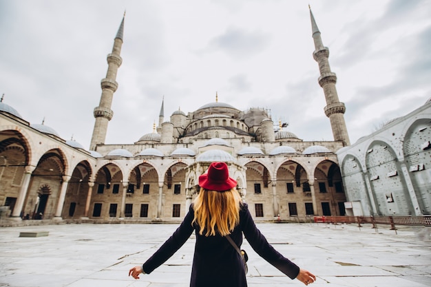 Joven viajero en sombrero y abrigo negro mirando la mezquita azul en Estambul, Turquía. Una niña camina durante el invierno en Estambul. Foto de rubia de viaje en el fondo de una mezquita en día de otoño.