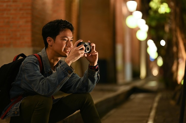 Joven viajero tomando fotos con su cámara sentado en las escaleras en la ciudad nocturna con fondo borroso de luces de la calle