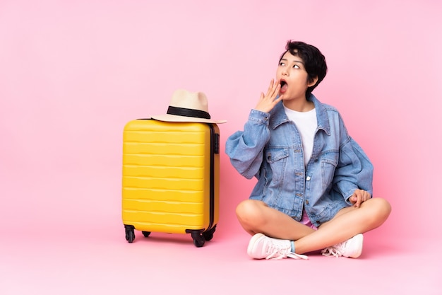 Joven viajero vietnamita con maleta sentada en el suelo sobre rosa aislado bostezando y cubriendo la boca abierta con la mano