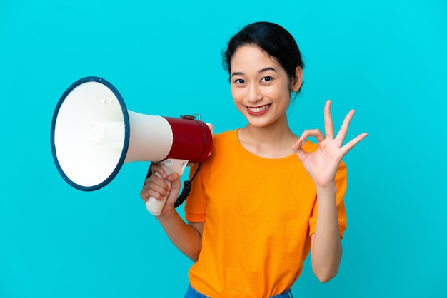 Joven vietnamita aislada de fondo azul sosteniendo un megáfono y mostrando el signo de ok con los dedos