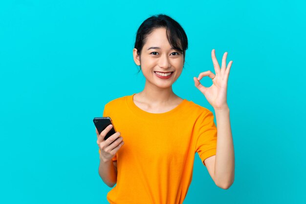 Joven vietnamita aislada de fondo azul usando teléfono móvil y haciendo el signo de OK