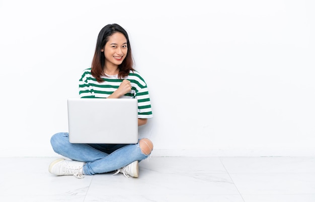 Joven vietnamita con una computadora portátil sentada en el suelo aislada en una pared blanca celebrando una victoria