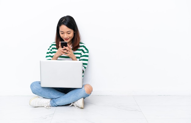 Joven vietnamita con una computadora portátil sentada en el suelo aislada en una pared blanca enviando un mensaje con el móvil