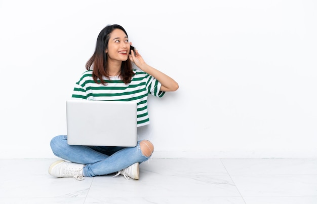 Joven vietnamita con una computadora portátil sentada en el suelo aislada en una pared blanca manteniendo una conversación con el teléfono móvil