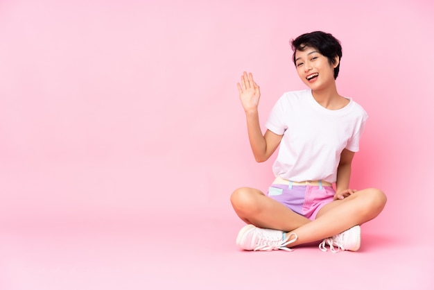 Joven vietnamita con el pelo corto sentada en el suelo sobre la pared rosa saludando con la mano con expresión feliz