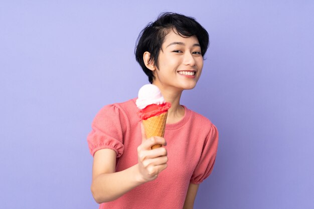 Joven vietnamita con el pelo corto sosteniendo un helado de cucurucho sobre la pared púrpura con expresión feliz