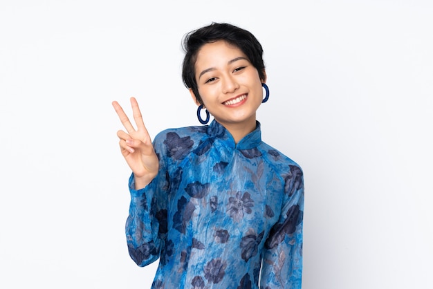Joven vietnamita con el pelo corto con un vestido tradicional sobre la pared blanca sonriendo y mostrando el signo de la victoria