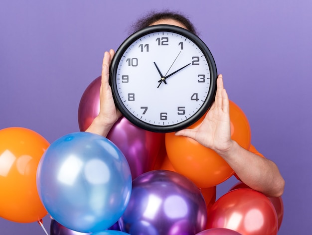 Joven vistiendo camiseta naranja de pie detrás de globos cubiertos de cara con reloj de pared