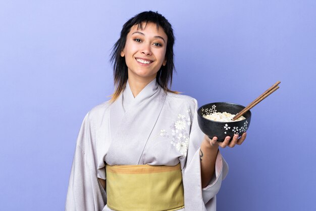 Joven vistiendo kimono sobre pared azul sonriendo mucho mientras sostiene un tazón de fideos con palillos