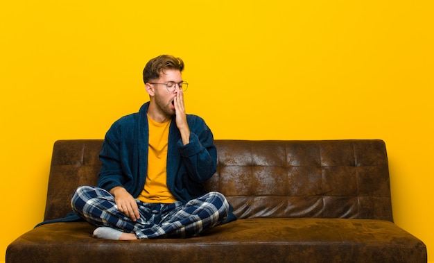 Joven vistiendo pijama bostezando perezosamente temprano en la mañana, despertando y luciendo somnoliento, cansado y aburrido. sentado en un sofa