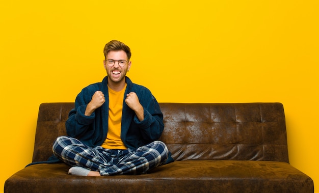 Joven vistiendo pijama gritando triunfante, riendo y sintiéndose feliz y emocionado mientras celebraba el éxito. sentado en un sofa