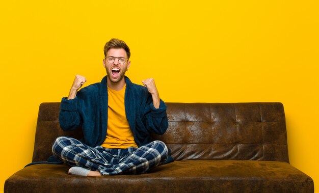 Joven vistiendo pijama sintiéndose feliz, positivo y exitoso, celebrando la victoria, los logros o la buena suerte. sentado en un sofa