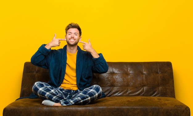 Joven vistiendo pijamas sonriendo con confianza apuntando a su propia sonrisa amplia, actitud positiva, relajada y satisfecha. sentado en un sofa