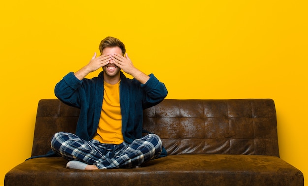 Joven vistiendo pijamas sonriendo y sintiéndose feliz, cubriendo los ojos con ambas manos y esperando una sorpresa increíble. sentado en un sofa