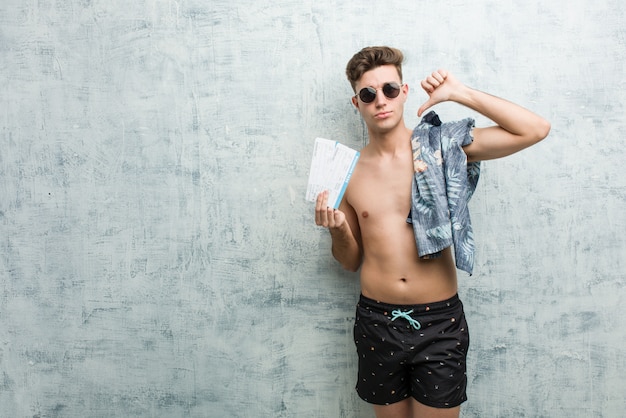 Joven vistiendo un traje de baño con billetes de avión mostrando un gesto de disgusto, pulgares hacia abajo. Concepto de desacuerdo