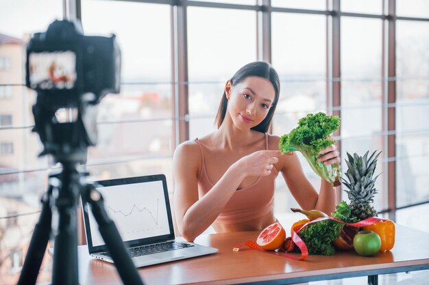 Joven vlogger de fitness haciendo videos en interiores sentado cerca de la mesa con comida saludable.