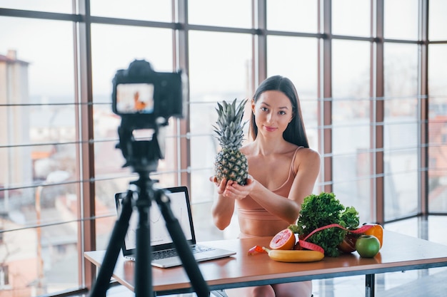 Joven vlogger de fitness haciendo videos en interiores sentado cerca de la mesa con comida saludable.