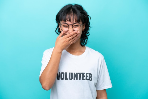 Joven voluntaria argentina aislada de fondo azul feliz y sonriente cubriendo la boca con la mano