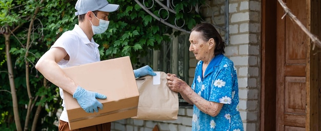 Un joven voluntario enmascarado le da a una anciana cajas con comida cerca de su casa. El hijo ayuda a una madre anciana soltera. Apoyo familiar, cuidado. En cuarentena, aislado. Coronavirus (COVID-19. Donación