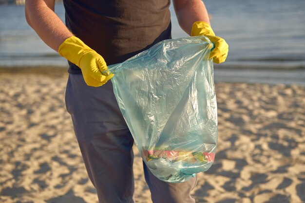 Joven voluntario en guantes amarillos está caminando con bolsa de basura a lo largo de una playa sucia del río