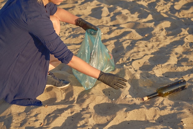 Joven voluntario con guantes negros está caminando con una bolsa de basura a lo largo de una playa sucia del río.