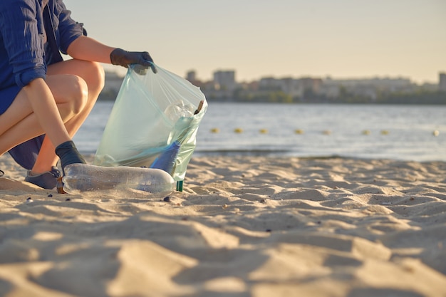 Joven voluntario con guantes negros está caminando con una bolsa de basura a lo largo de una playa sucia del río.