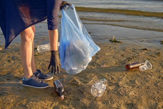 Joven voluntario con guantes negros está caminando con una bolsa de basura a lo largo de una playa sucia del río.