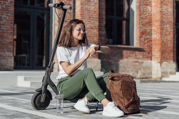 Una joven con zapatillas blancas está sentada en un scooter eléctrico y mirando un reloj inteligente