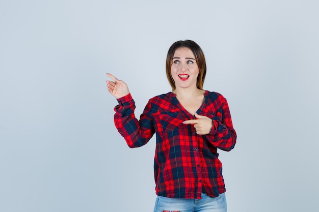 Jovencita apuntando hacia el lado izquierdo, abriendo la boca con camisa a cuadros, jeans y mirando sorprendido, vista frontal.