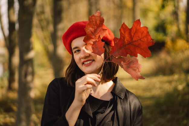 Una jovencita en una boina roja con un ramo de hojas de otoño en sus manos camina por el bosque