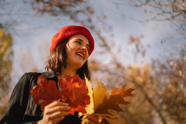Una jovencita en una boina roja con un ramo de hojas de otoño en sus manos camina por el bosque