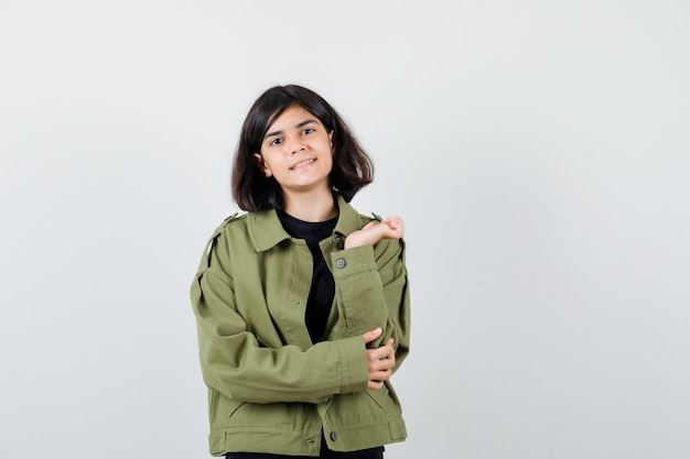 Jovencita en camiseta, chaqueta verde extendiendo la palma a un lado mientras mira a la cámara y mira alegre, vista frontal.