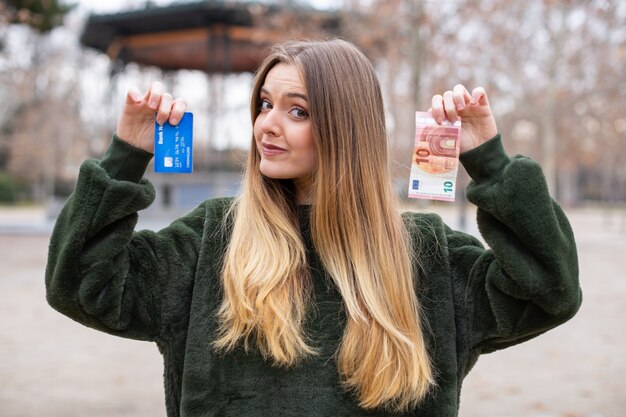 Jovencita demostrando tarjeta de plástico y dinero en efectivo a la cámara mientras está de pie en el parque