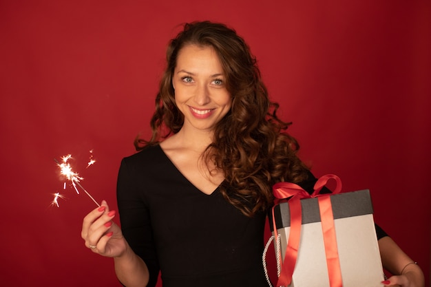 Jovencita encantadora con caja de regalo y bengala ardiente sonriendo mirando a cámara sobre fondo rojo. Regalos de fiesta de Navidad, celebración de Nochevieja, concepto de ideas para regalar. Copia espacio