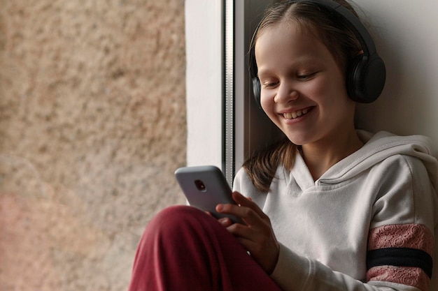 Jovencita escuchando música de audio de audiolibros con auriculares y móvil en la ventana