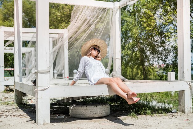 Jovencita glamorosa disfruta del estilo de vida y posa contra un cenador de madera blanca cerca de la playa del lago