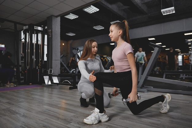 Jovencita haciendo estocadas con pesas trabajando con entrenador personal en el gimnasio