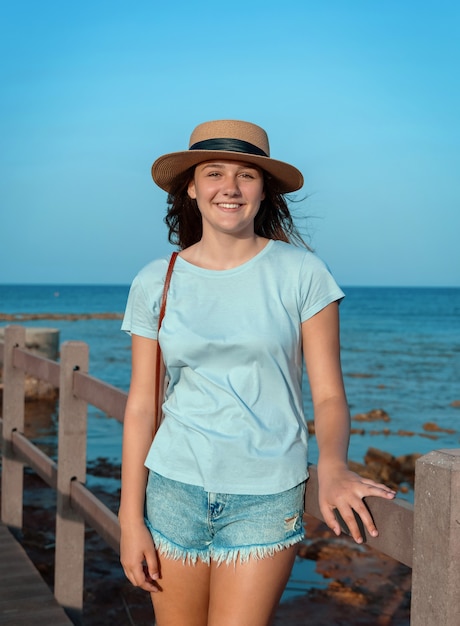 Jovencita de pie sobre la pasarela de madera junto al mar al atardecer, vestida con camiseta azul claro, sombrero de paja y bolso. Concepto de viaje de verano