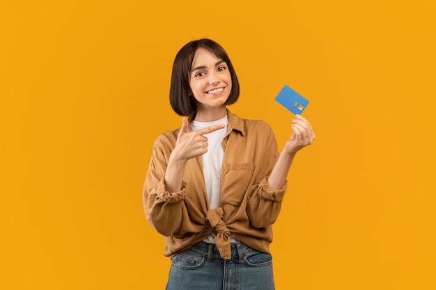 Jovencita positiva señalando la tarjeta de crédito recomendando el servicio bancario posando sobre fondo amarillo y sonriendo